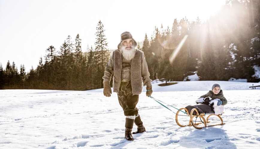 Älterer Mann zieht ein lachendes Kind auf einem Holzschlitten über den Schnee, im Hintergrund Wald und tief stehende Wintersonne.