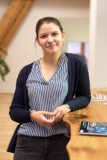 Junge Frau in gestreifter Bluse lehnt lächelnd an einer Holztheke im Büro. Daneben liegen Magazine.