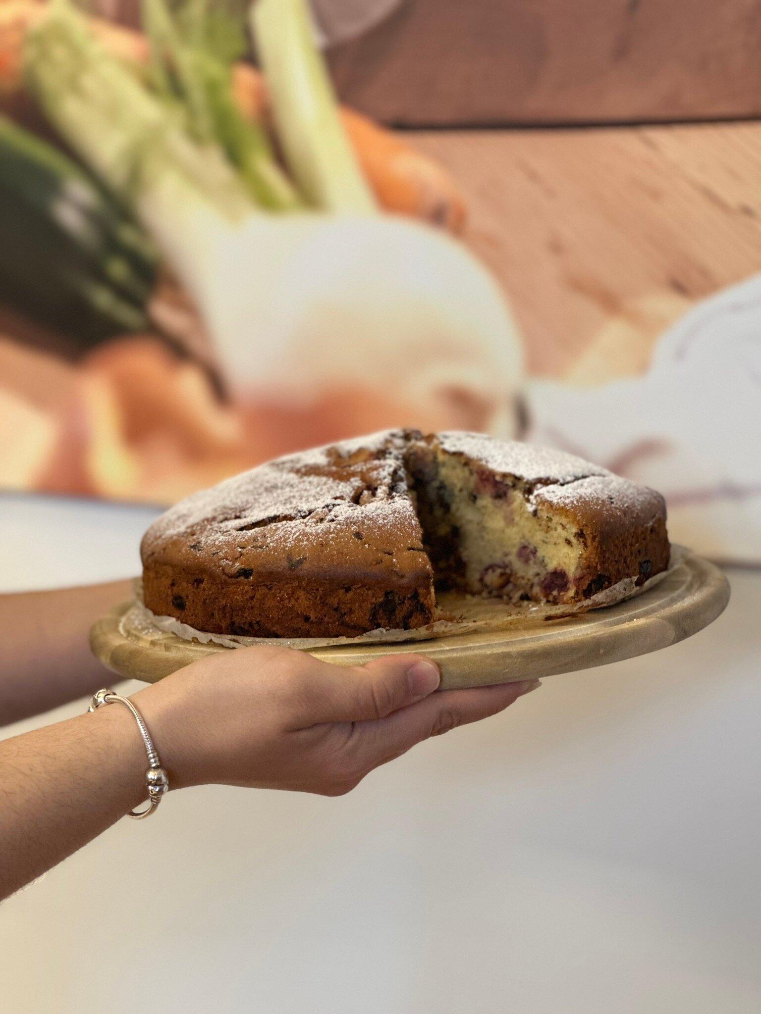 Hand hält angeschnittenen Kirschkuchen, mit Staubzucker bestäubt, auf Holzbrett vor unscharfem Gemüsehintergrund.