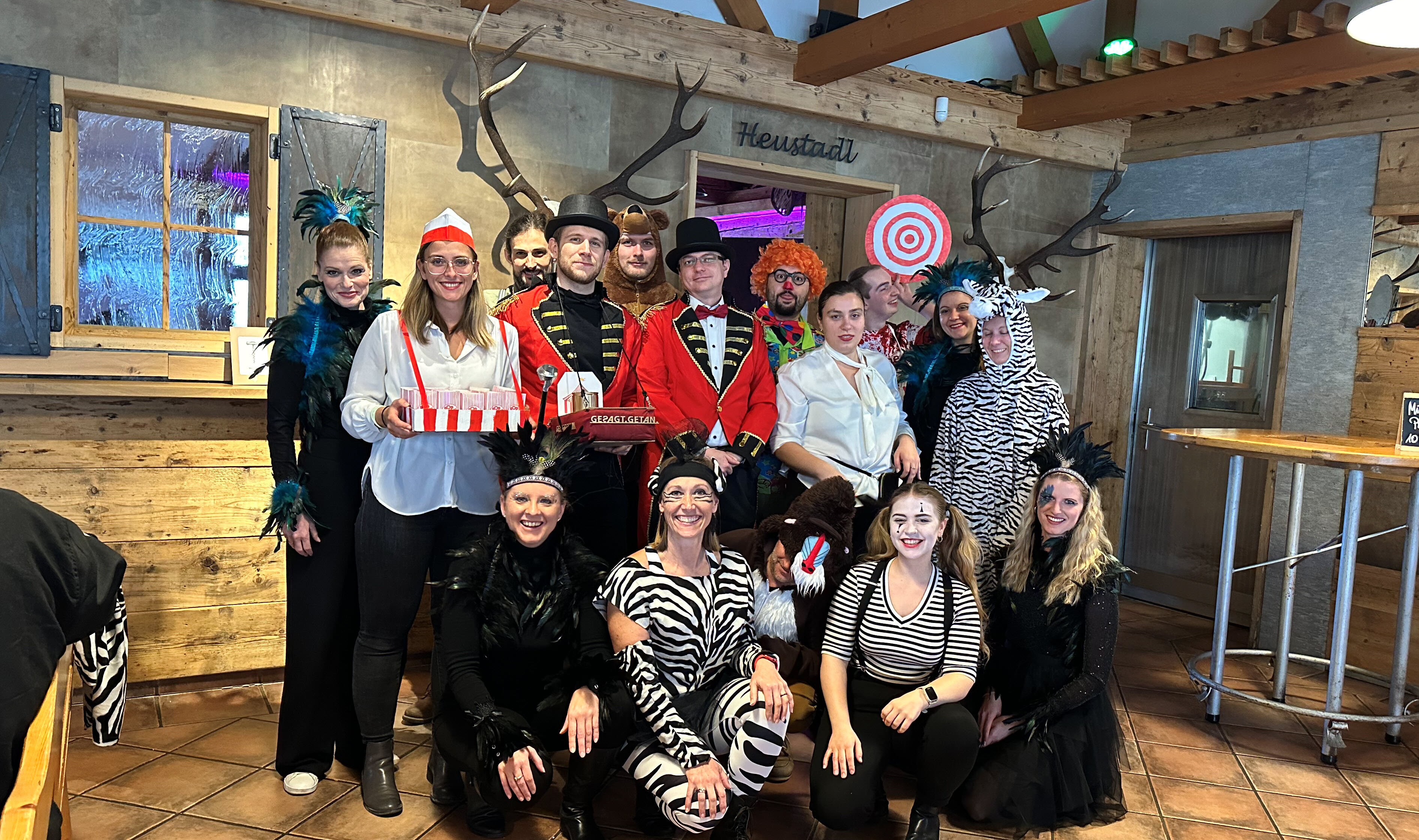 Ein Team in Zirkus- und Tierkostümen posiert im rustikalen Raum mit Dompteur, Clown, Zebras und Hirschen, passend zur Faschingsfeier.